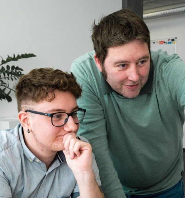 Der KV-Lernende sitzt am Computer, während sein Berufsbildner ihm etwas zeigt.